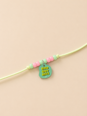 A light green rakhi for dogs and cats featuring pastel pink and mint green beads, with a mint green guitar-shaped charm displaying the text 'BRO GOT RIZZ' in yellow.