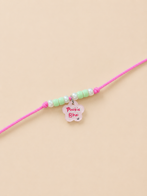 A pink rakhi for dogs and cats for featuring pastel green and white beads, a small white flower charm with pink text reading 'Pookie Bhai', and a silver ring connector, displayed on a solid beige background.
