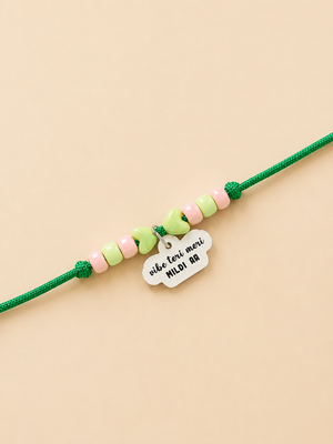 A green pet rakhi featuring pastel pink and light green beads, a mint green bow charm, and a white cloud-shaped tag engraved with 'vibe teri meri mildi aa'.