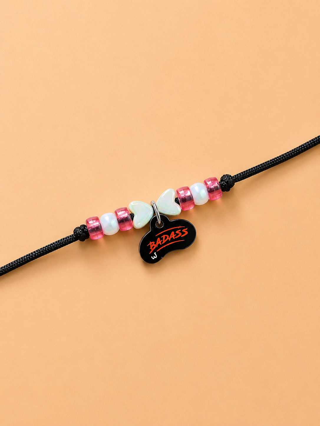 A black dog rakhi featuring a central charm shaped like a black bean with the word 'BADASS' written in red, flanked by pink and white beads on a peach-colored background.