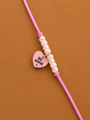 A pink dog rakhi for pets featuring a heart-shaped charm with 'BE MINE' printed in black, accented by light pink and white beads on a solid brown background.