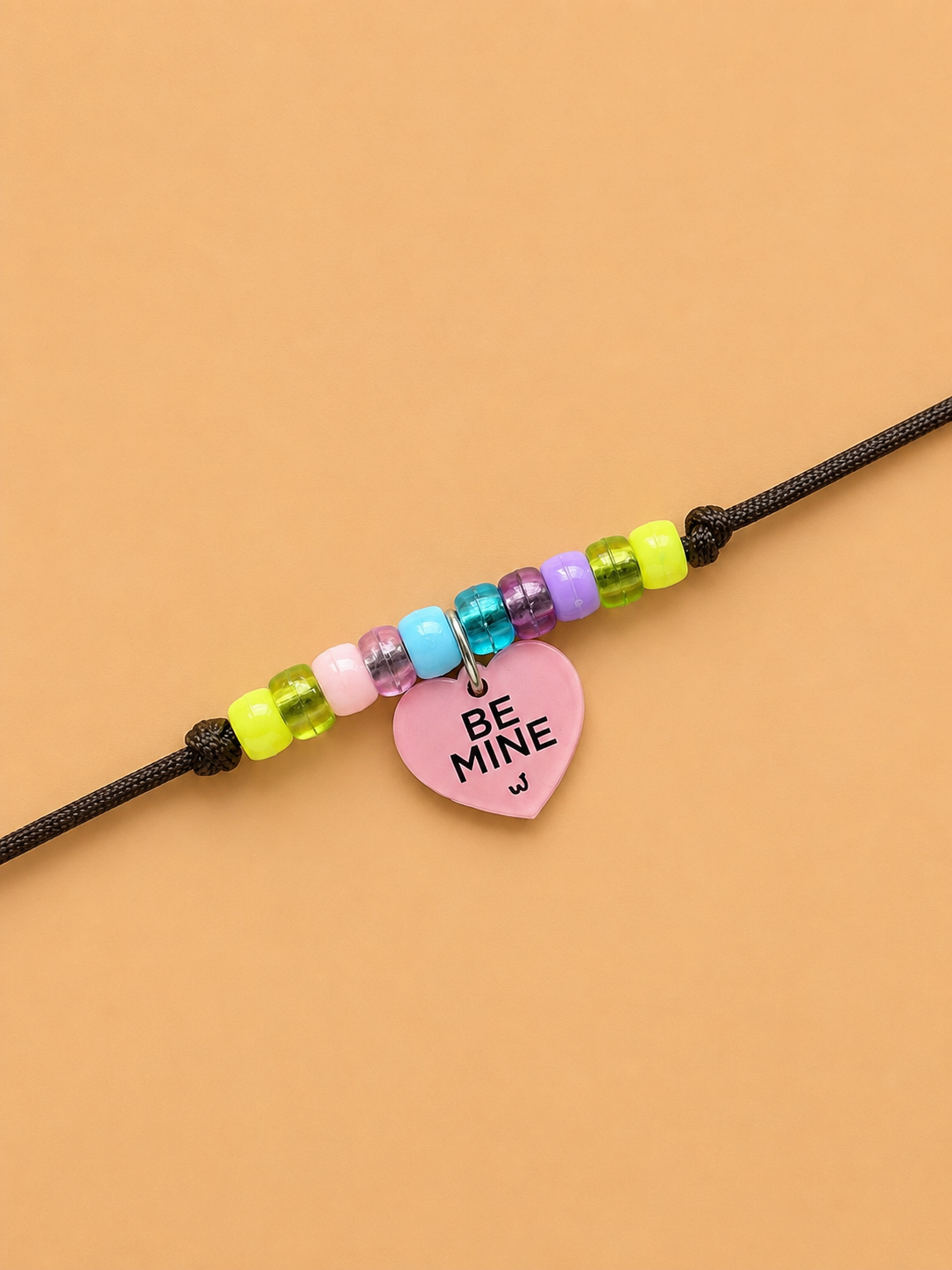 rakhi for dogs and cats with a pink heart-shaped charm that reads 'BE MINE' in black text, displayed on a solid peach background.