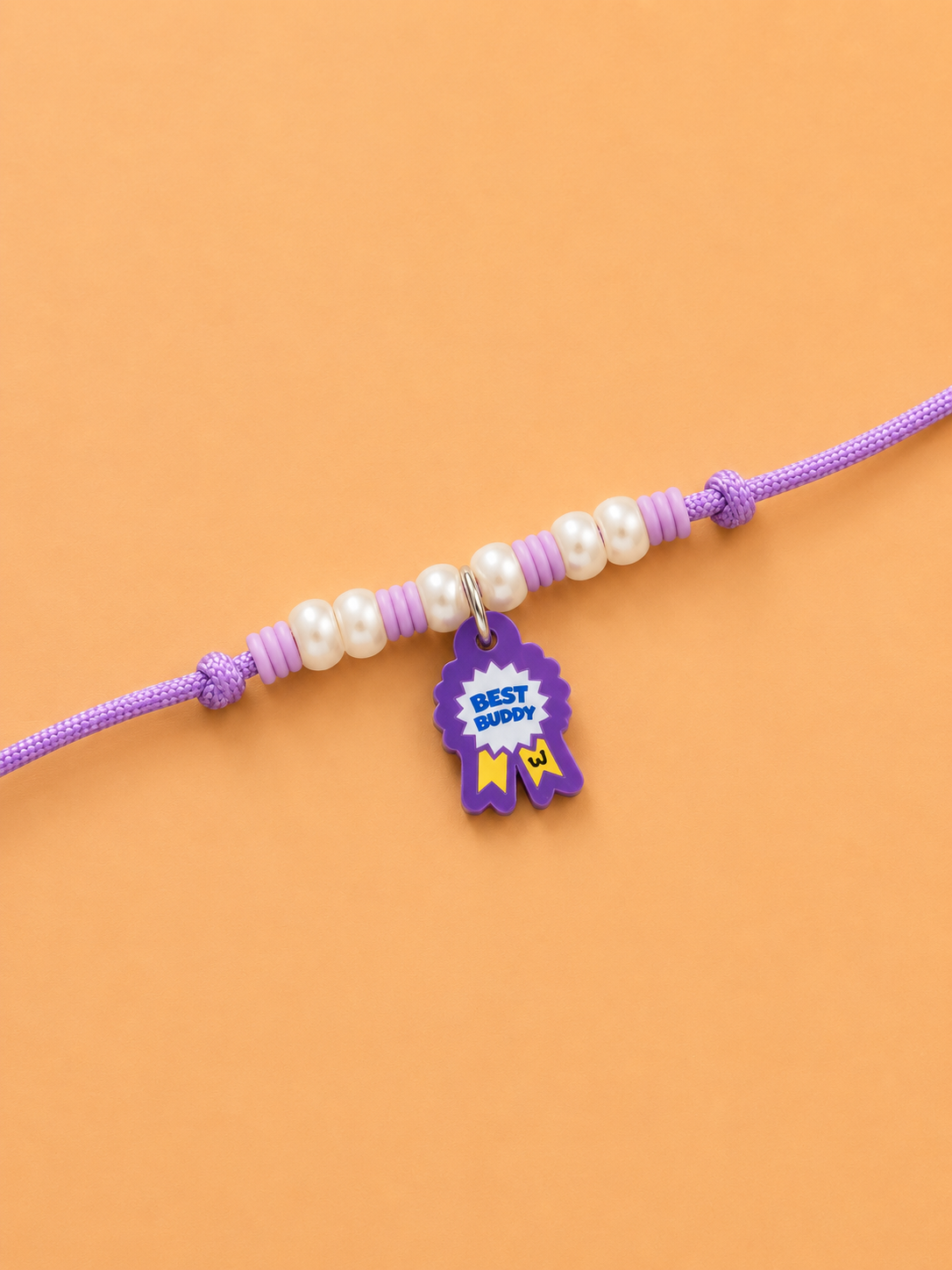A purple rakhi for dogs and cat featuring white faux pearls, purple spacer beads, and a small purple ribbon award charm with 'BEST GIFTY' written on it, displayed on a solid orange background.