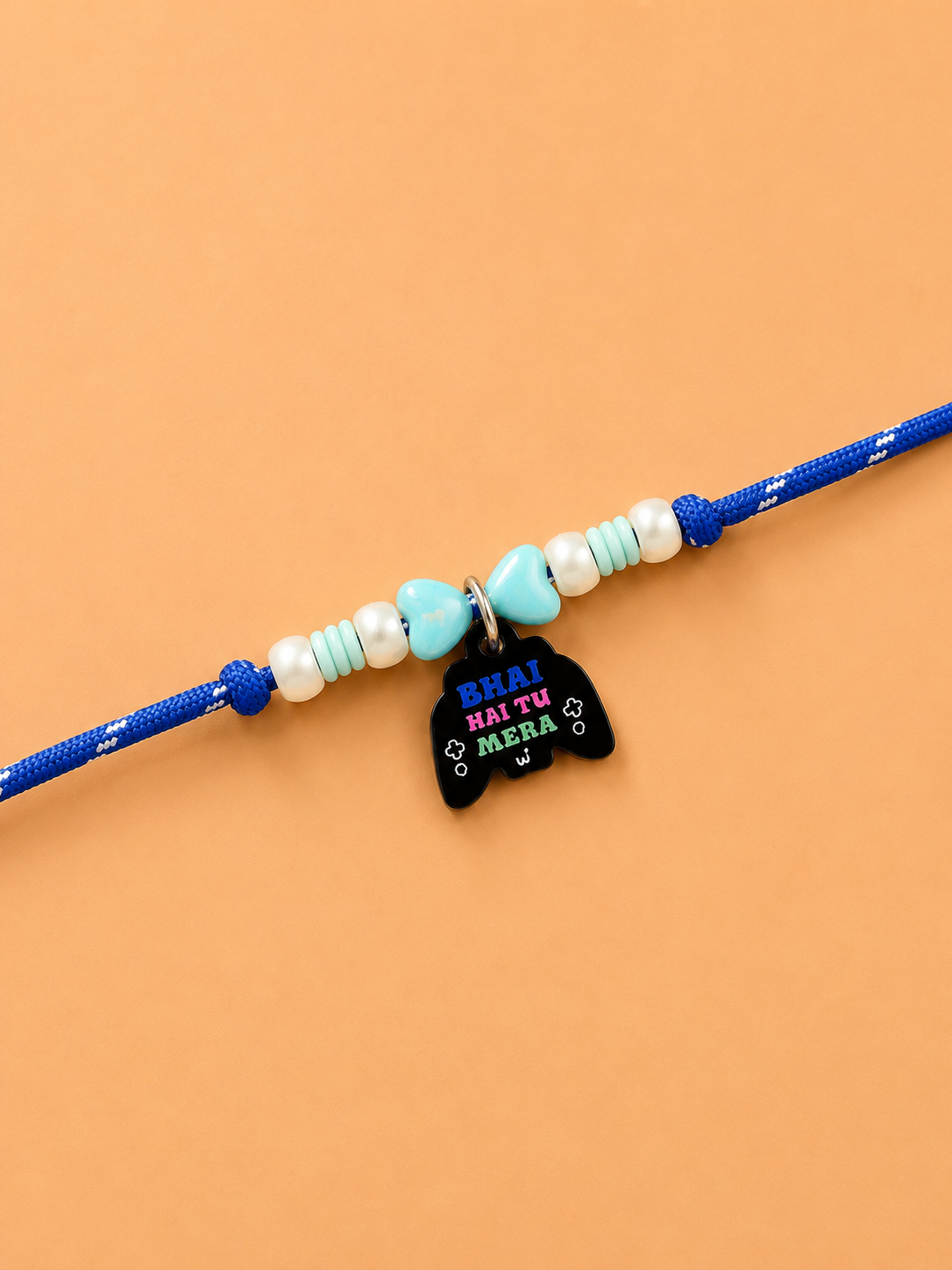 A blue dog pet rakhi featuring white and light blue beads, a small turquoise heart, and a black cat-shaped charm with the text 'HEAI HAI TU MERA' in colorful lettering, displayed on a solid peach background.