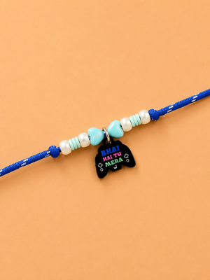 A blue dog pet rakhi featuring white and light blue beads, a small turquoise heart, and a black cat-shaped charm with the text 'HEAI HAI TU MERA' in colorful lettering, displayed on a solid peach background.