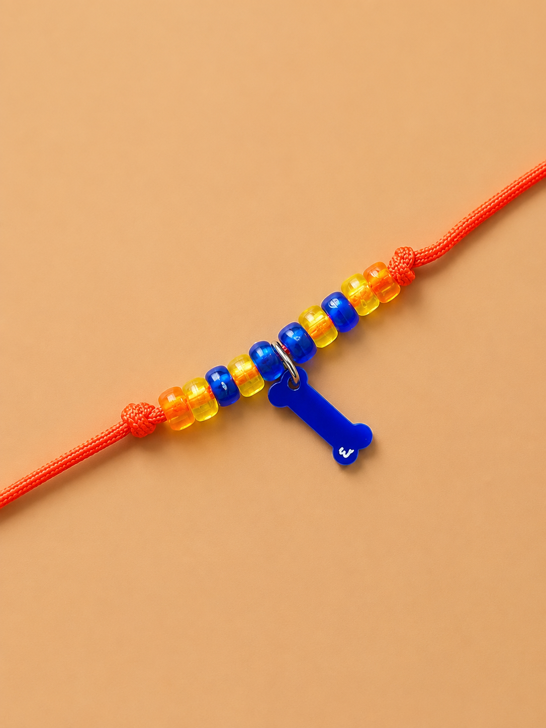 rakhi for dogs and cats featuring translucent yellow and blue beads, with a blue plastic letter 'I' charm, set against a solid tan background.