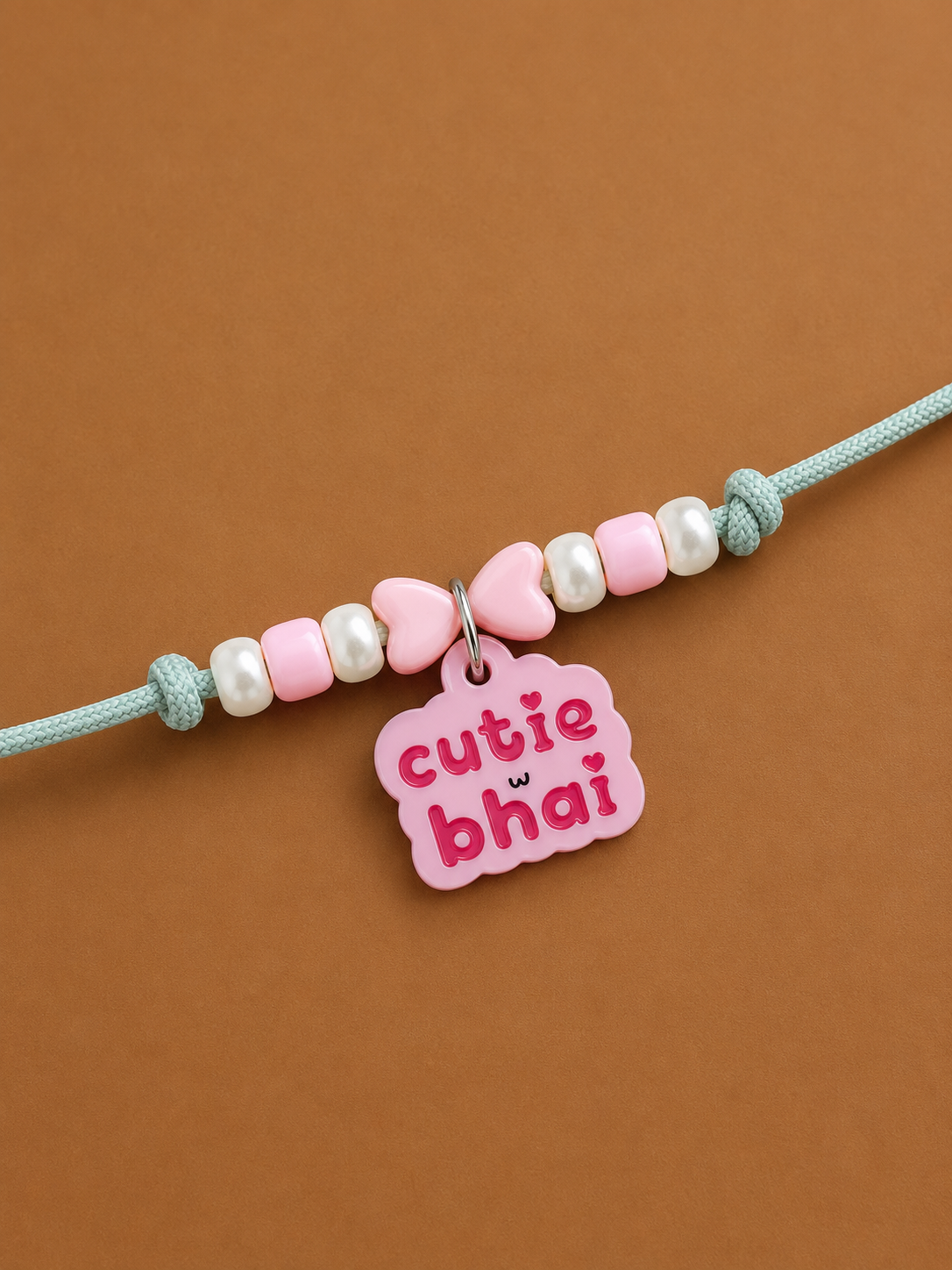 A grey dog paw rakhi featuring pink and white beads, two pink heart charms, and a pink tag that reads 'cutie bhai' in pink lettering, displayed on a solid brown background.