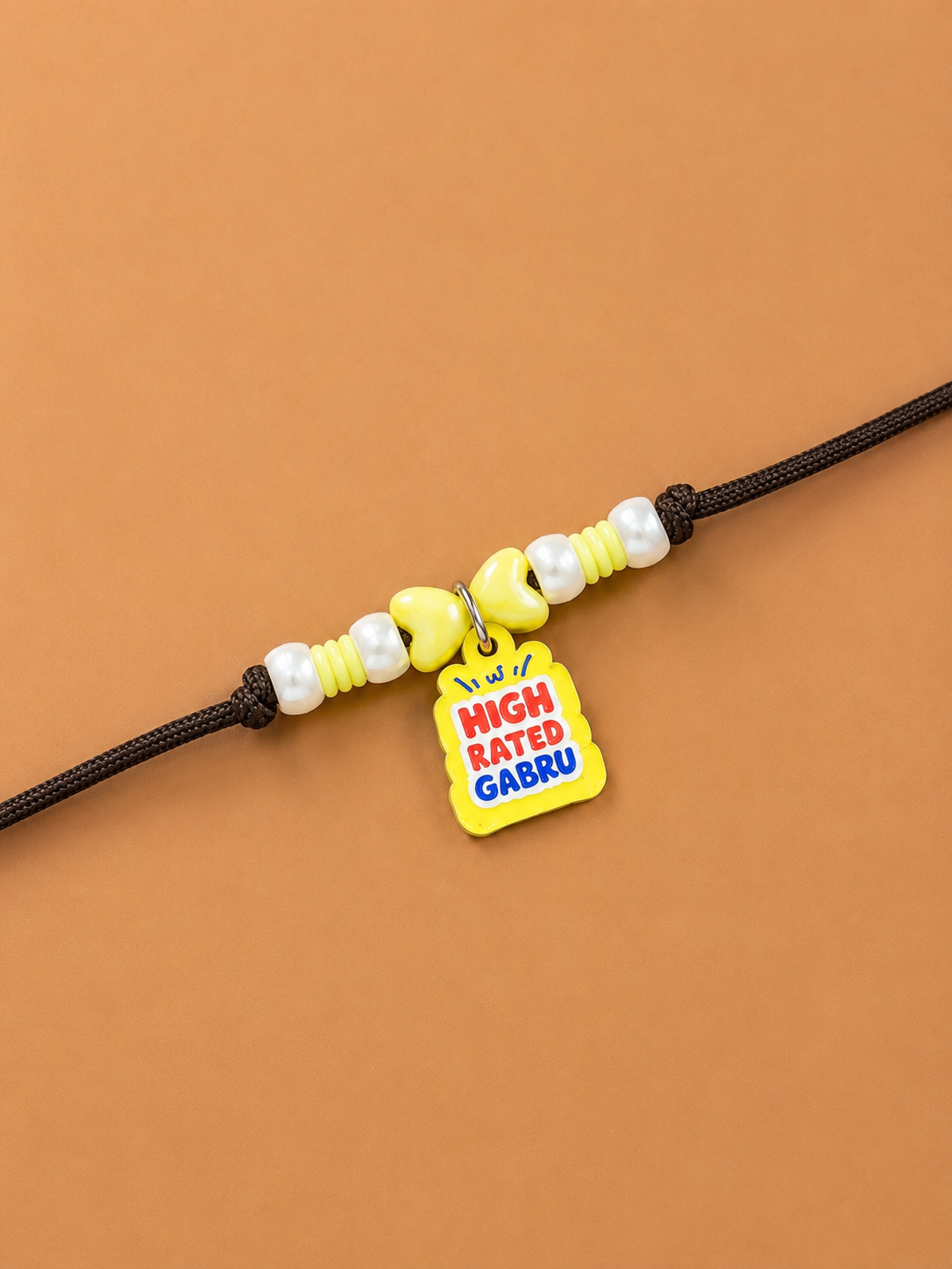 A brown rakhi for dogs and cats  featuring yellow and white beads, a small yellow bow charm, and a yellow tag charm printed with the text 'I love HIGH RATED GABRU' in red and blue, set against a solid warm brown background.