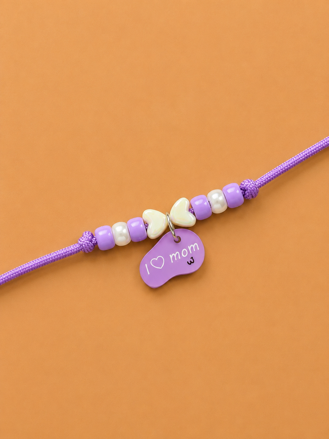 A purple dog paw rakhi with purple and cream beads, a small cream bow charm, and a purple heart-shaped tag engraved with 'I love mom'.