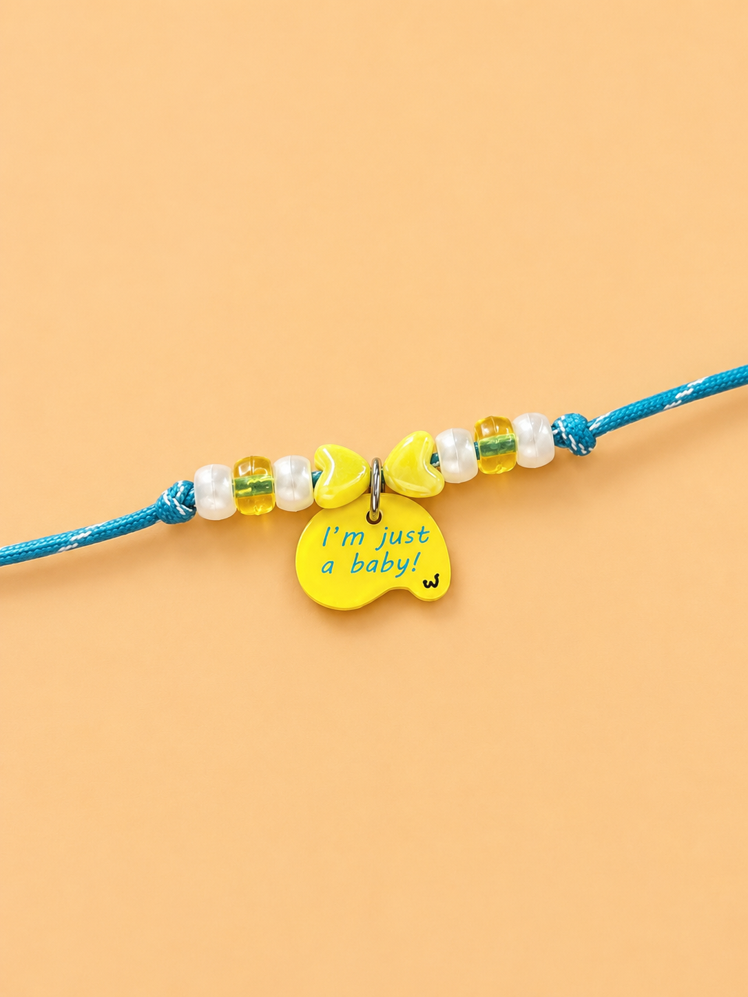 A teal rakhi for pets featuring white faux pearls, yellow beads, two yellow heart charms, and a central yellow apple charm with the text 'I'm just a baby!' in blue lettering, displayed on a solid peach background.