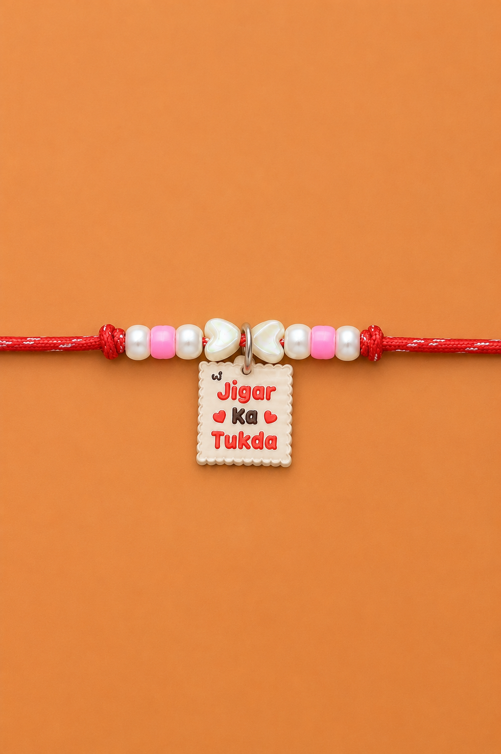 A red pet rakhi for dogs and cats with pink and white beads, two white pearl-like beads, and a small white rectangular charm engraved with 'Jigar Ka Tukda' in red text, shown on an orange background.
