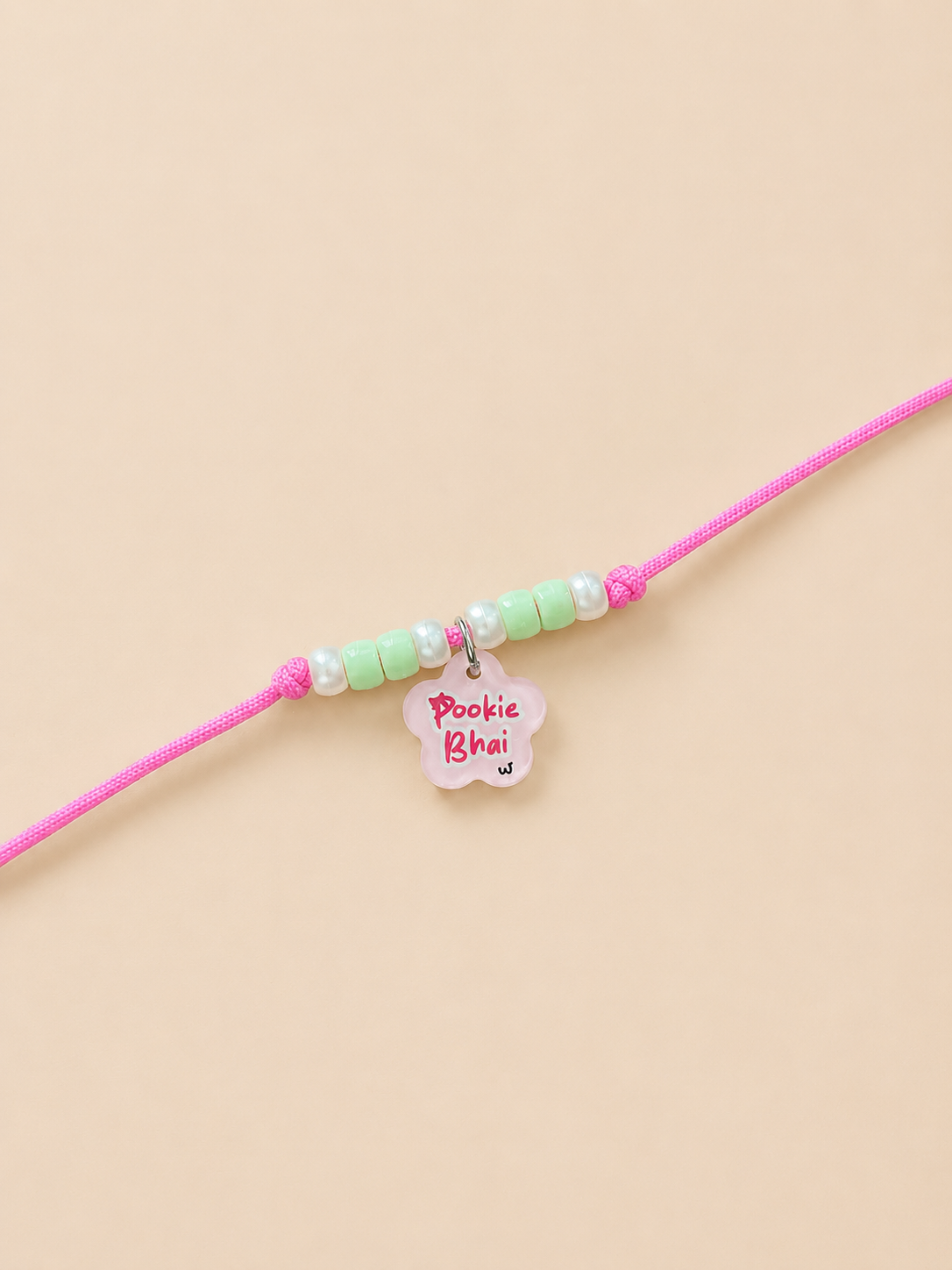A pink rakhi for dogs and cats for  featuring pastel green and white beads, a small white flower charm with pink text reading 'Pookie Bhai', and a silver ring connector, displayed on a solid beige background.