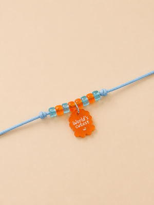 A light blue dog rakhi featuring a row of alternating light blue and orange beads, with a small orange tag hanging from the center that reads 'World's cutest'.