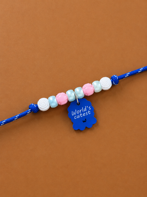 A blue rakhi for dogs and cats with pastel white, light blue, pink, and white beads, featuring a blue scalloped charm that reads 'World's cutest' in white text, displayed on a solid brown background.
